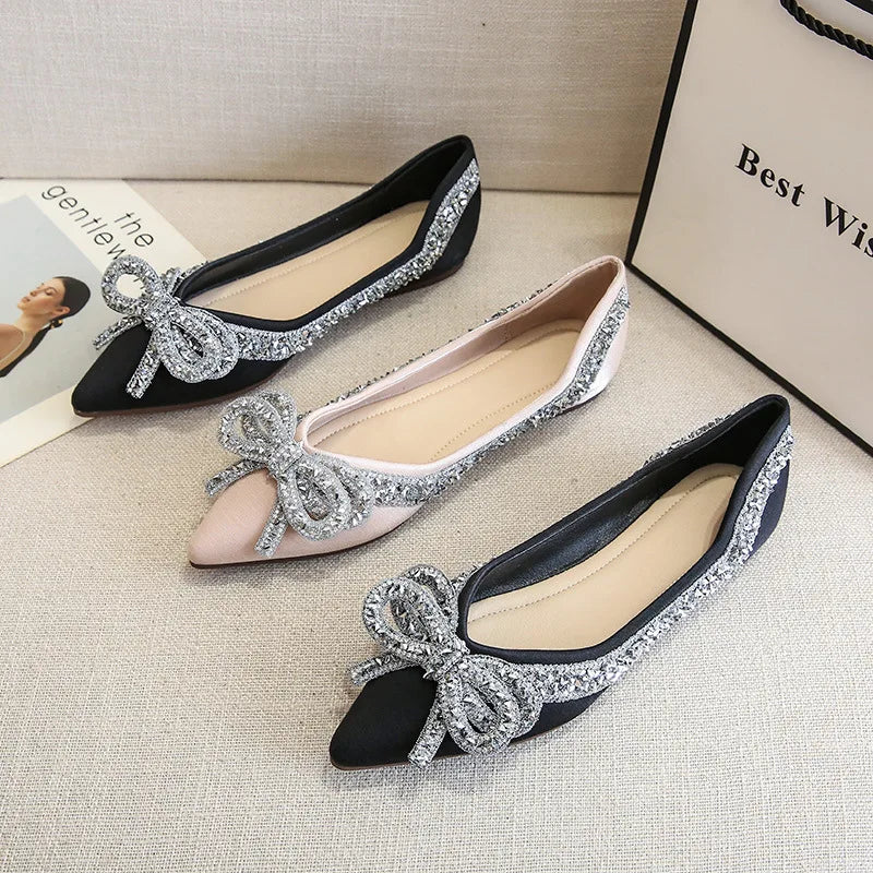 Three embellished women's flats, two black and one beige, featuring sparkly bows and trim, placed on a beige surface with a magazine and gift bag nearby.