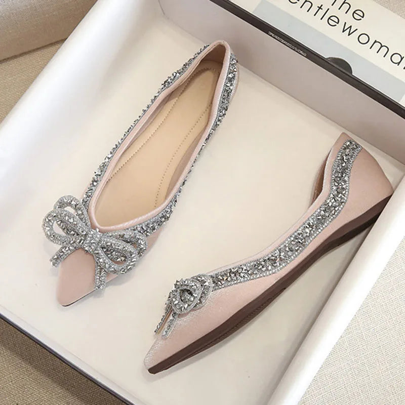 Elegant pink ballet flats with sparkling silver rhinestone bows and trim, placed in a white shoebox on a beige surface.