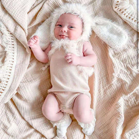 Smiling baby wearing a fluffy bunny hat and beige onesie, lying on a soft cream-colored blanket with crocheted edges. The baby is wearing white lace socks, appearing cozy and playful.
