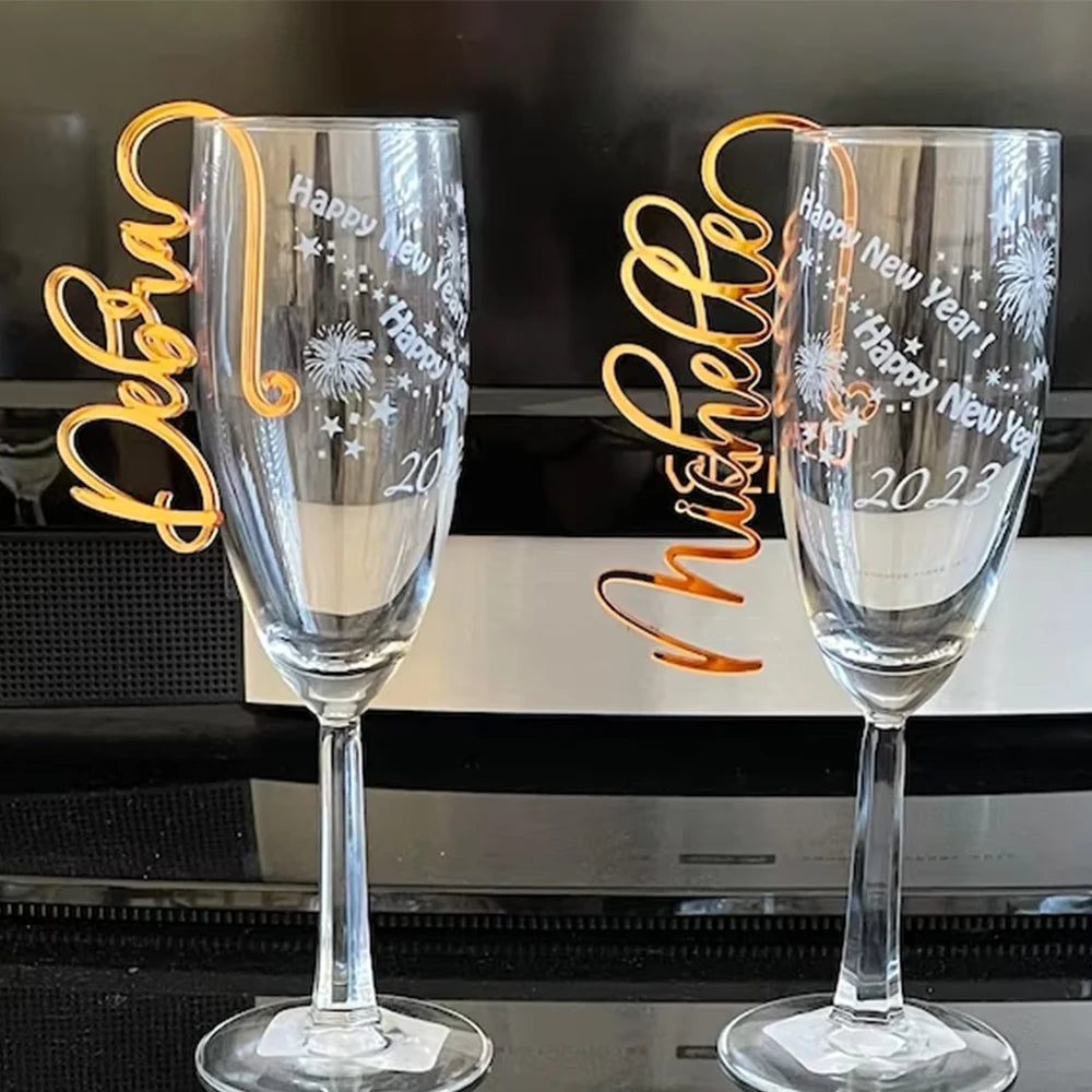Two champagne flutes etched with "Happy New Year 2023" and fireworks designs. Each has an orange name tag curling around the rim, one reading "Michelle" and the other "Deborah."