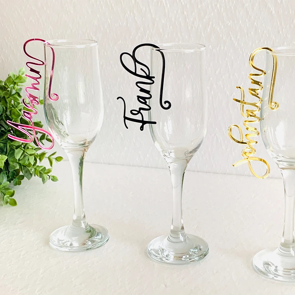 Three champagne flutes with cursive name tags in pink, black, and gold affixed to the rims. A green plant decorates the background.
