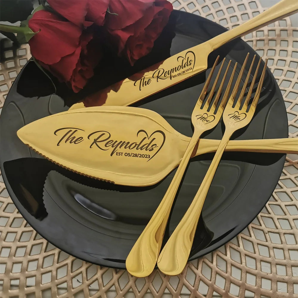 Gold cake serving set with "The Reynolds" and a date engraved, resting on a black plate. The set includes a knife and forks labeled "Mr." and "Mrs." with red roses nearby.