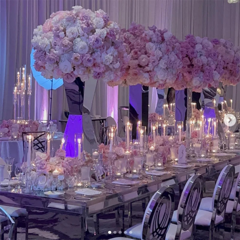 Elegant wedding reception with tall pink and white floral arrangements, mirrored tables, and candlelight. Plush chairs and soft purple lighting create a luxurious atmosphere.