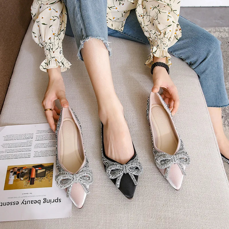 A person in a floral blouse and jeans tries on glittery pointed-toe flats with large bows, one shoe black and the other pink, while sitting on a cushioned bench next to an open magazine.