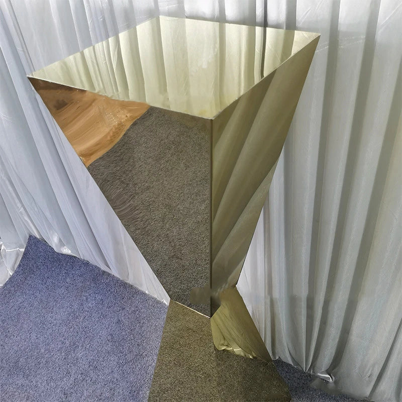 A shiny, brass-finished pedestal with a modern, angular design stands on a grey carpeted floor against a backdrop of white curtains.