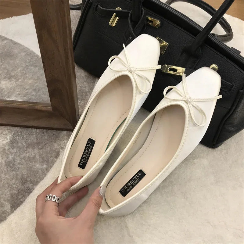 White ballet flats with bow detail, held by a hand with a silver ring, placed in front of a black handbag on a soft, light-colored surface.