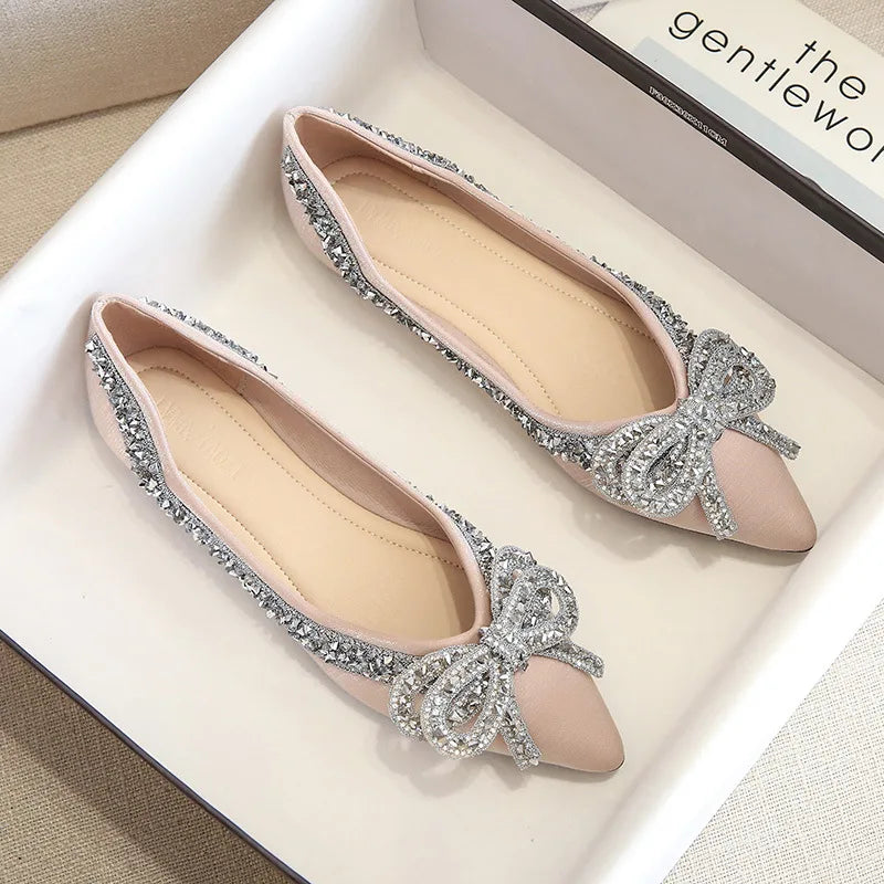 Elegant pink flats in a box, adorned with sparkling silver embellishments and bows on pointed toes, with a magazine partially visible in the background.