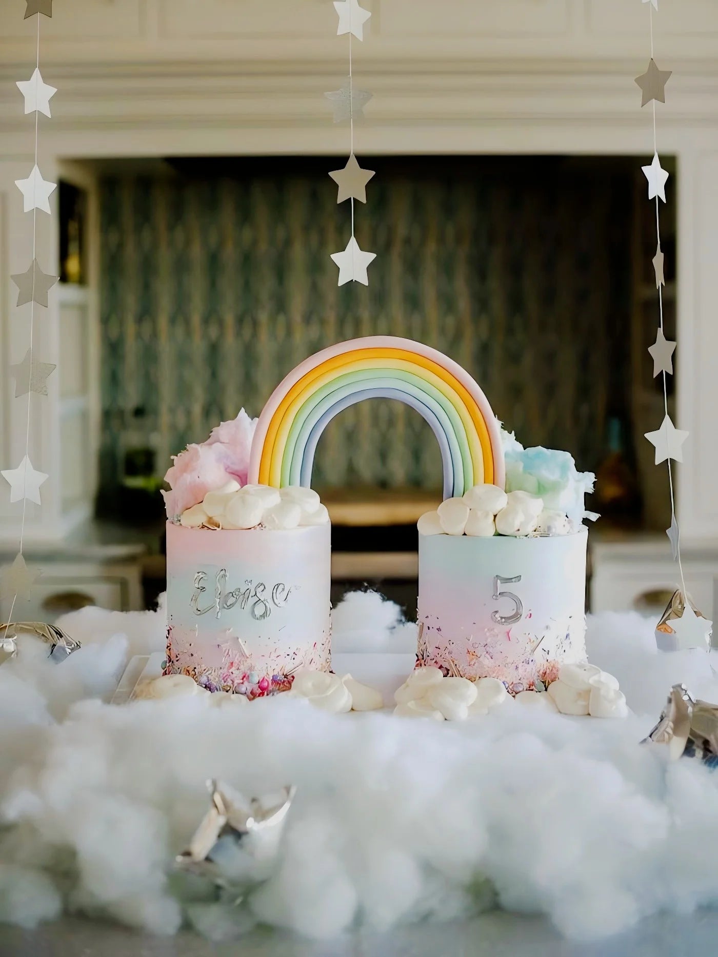 Two pastel cakes topped with a rainbow and clouds are surrounded by cotton-like fluff. Hanging star decorations add a whimsical touch. One cake reads "Eloise," the other "5."