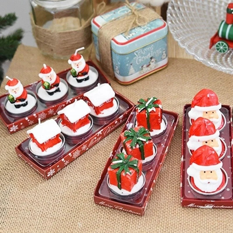 Festive candles shaped like Santa, gift boxes, and snow-covered houses, displayed in holiday-themed trays on burlap. A "Season's Greetings" tin and a decorative train are in the background.