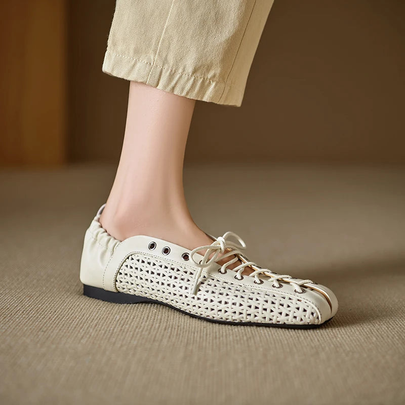 A person wearing beige pants is shown with a foot in a stylish white woven sneaker featuring laces and a black sole, on a beige carpeted floor.