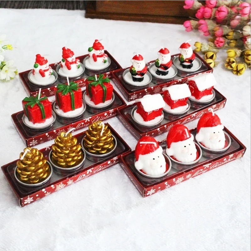 Festive Christmas candles shaped like Santa, gift boxes, gold pinecones, houses, and snowmen arranged in decorative packaging on a white surface, with pink flowers nearby.