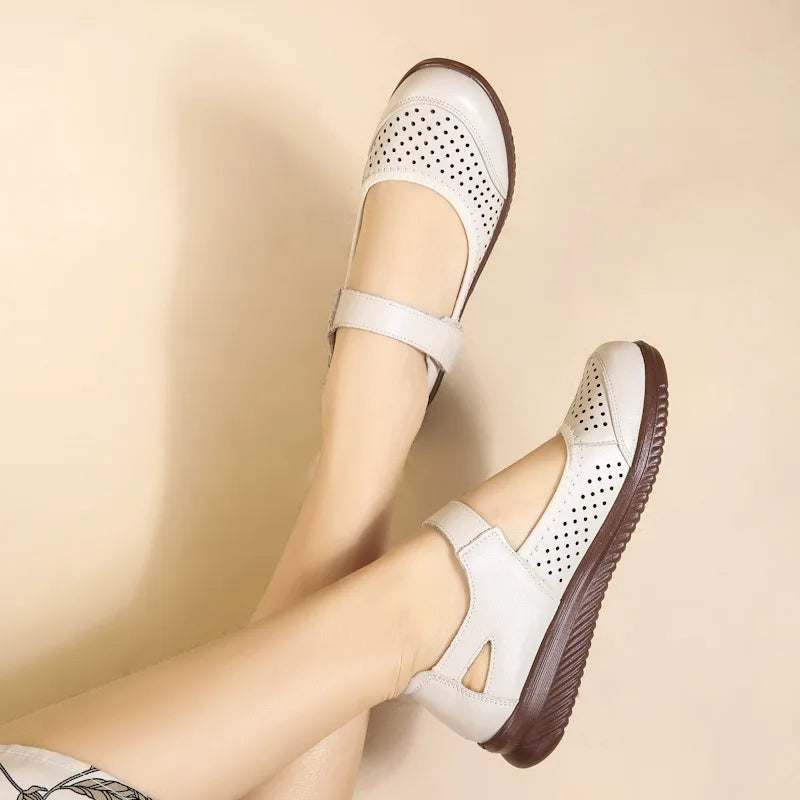 White Mary Jane shoes with perforated patterns and velcro straps, showcased on crossed legs against a beige background. The soles are a contrasting dark brown.