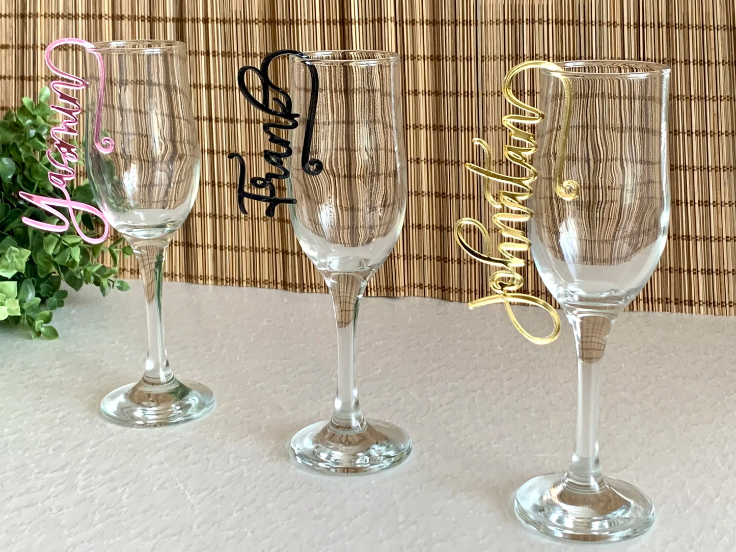 Three wine glasses with colored name tags—pink, black, and gold—displayed on a white surface in front of a bamboo backdrop, with green foliage on the left side.