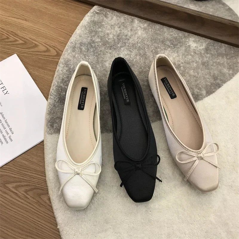 Three ballet flats in white, black, and beige with bow accents, arranged on a round rug near a wooden floor and an open book.