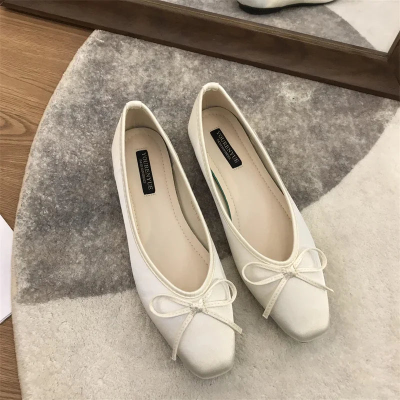 Elegant white ballet flats with squared toes and bow accents, placed on a circular gray and beige rug near a mirror reflecting part of the shoes.