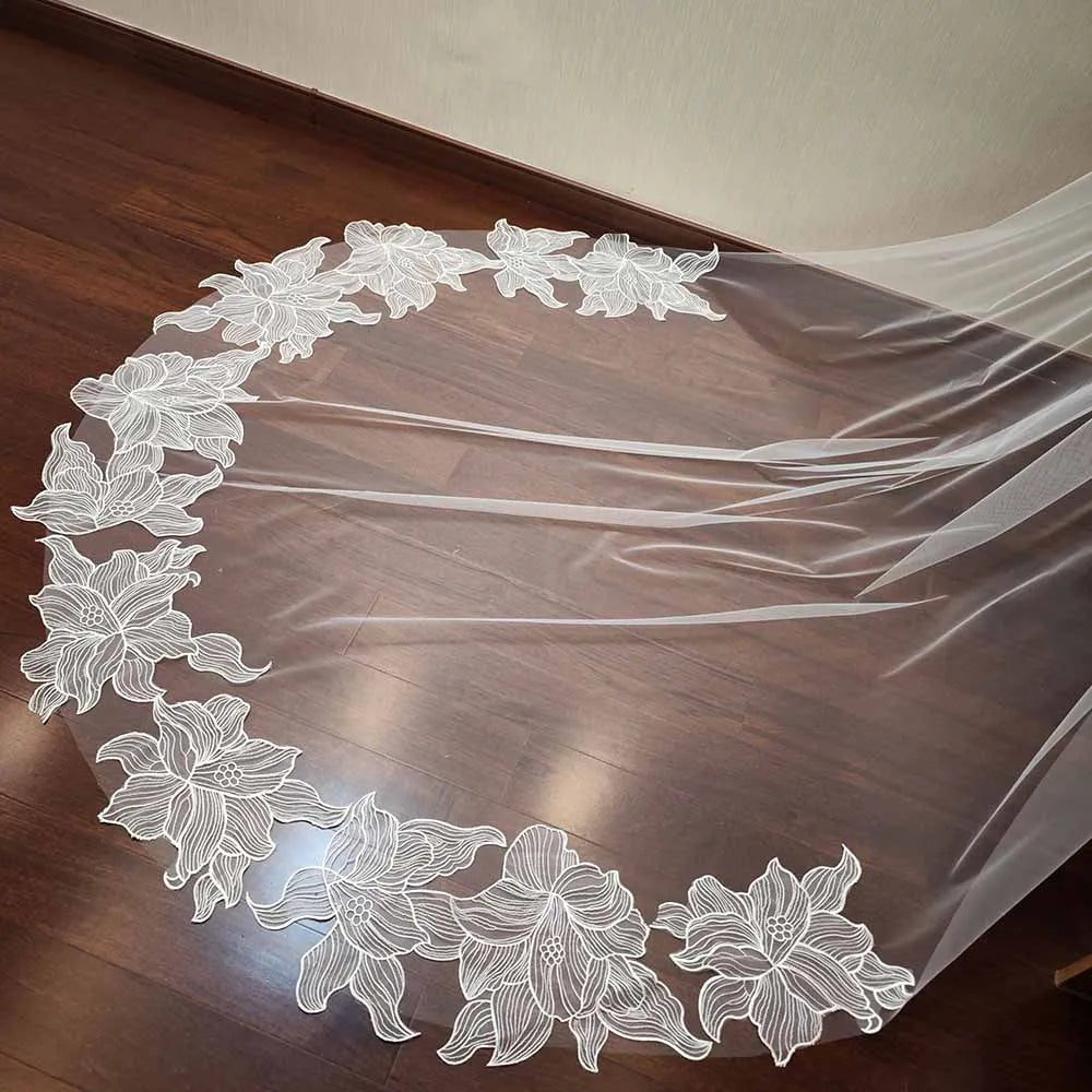 A sheer wedding veil with intricate white floral embroidery along the edge is spread out on a polished wooden floor.