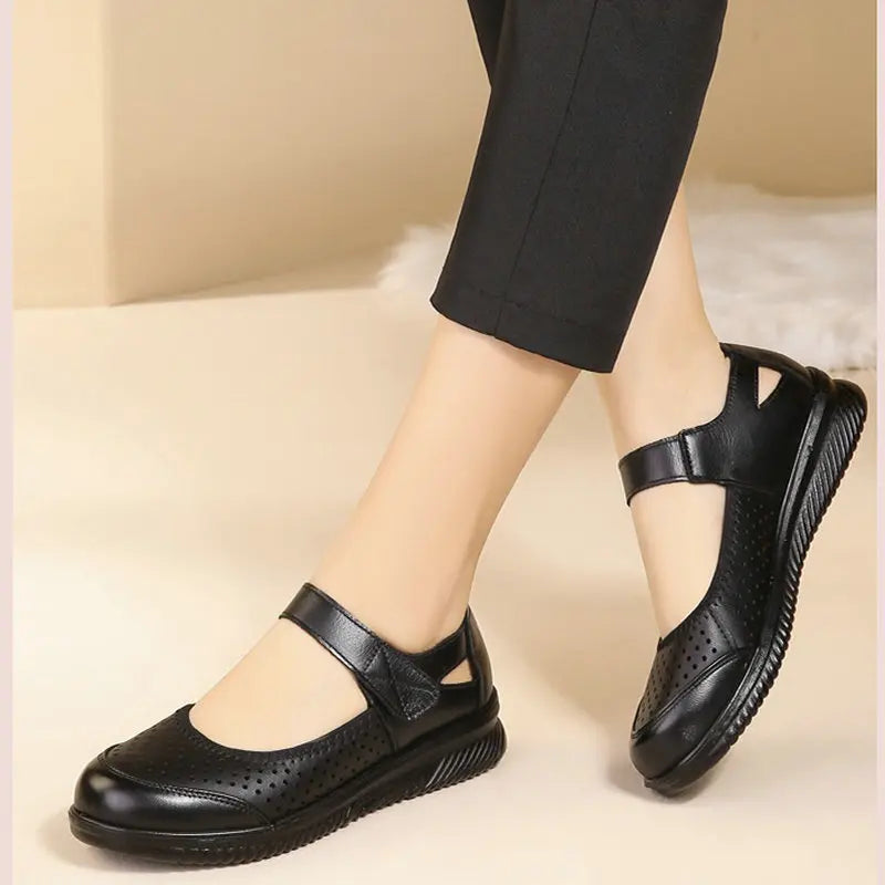 Person wearing black Mary Jane shoes with perforated design and ankle strap, paired with cropped black pants, standing on a light-colored floor.