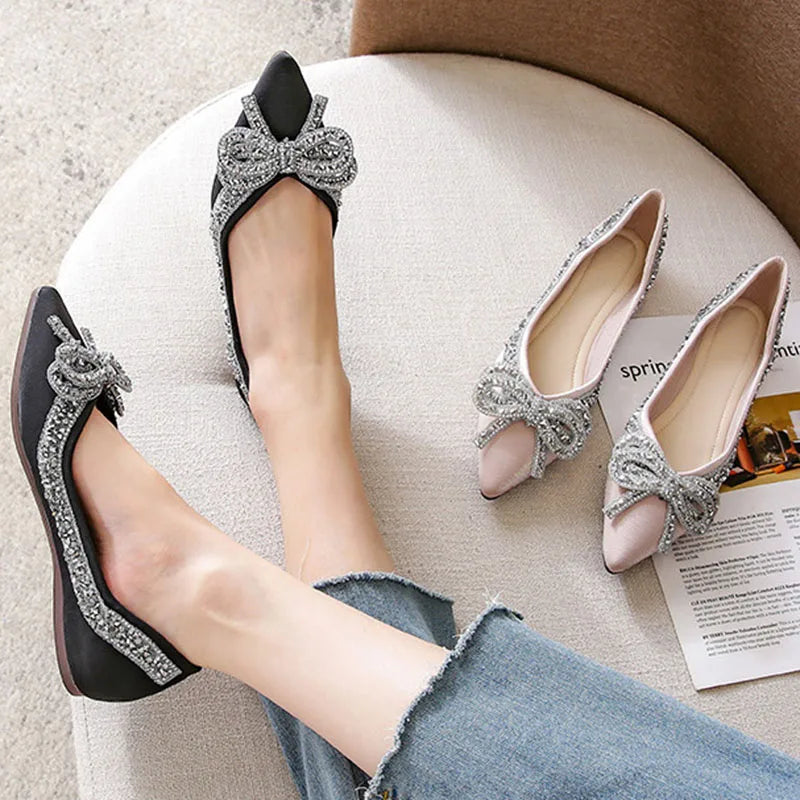 A person wearing black pointed flats adorned with silver rhinestone bows, sitting on a beige chair. Nearby are similar pink flats with the same decorative bows.
