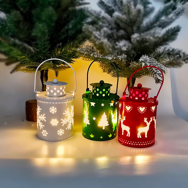 Three festive lanterns—white with snowflakes, green with trees, and red with reindeer—glow softly under small, snow-tipped evergreen trees on a wintry-themed tabletop.