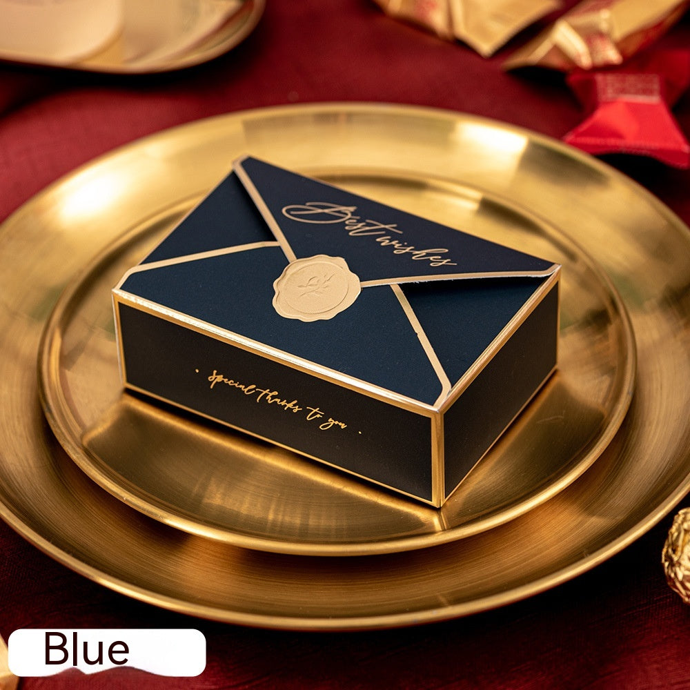 A small blue gift box with gold accents and "Best wishes" text on the lid, placed on a golden plate. The box has an envelope design with a wax seal detail.
