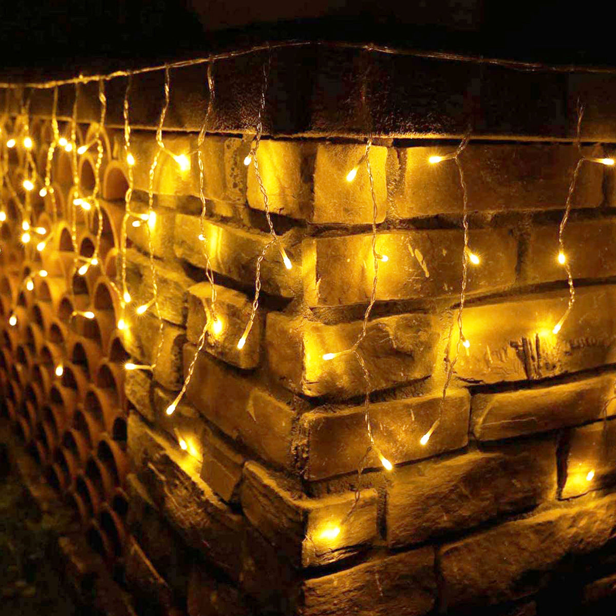 Warm white string lights drape over a rustic stone wall corner, illuminating the textured surface. The lights create a cozy and inviting nighttime ambiance.