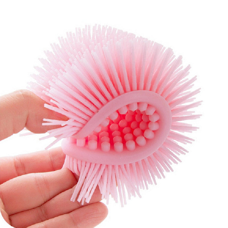 A hand holding a pink silicone scrubber with spiky bristles on one side and rounded nubs on the other, designed for cleaning or exfoliating.