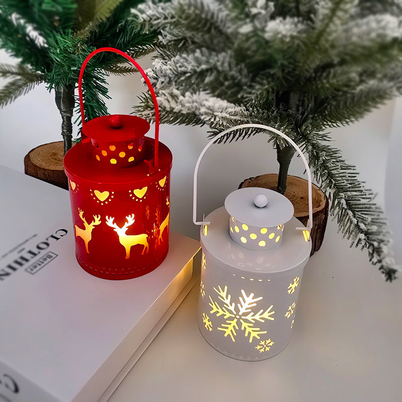 Red lantern with deer and heart cutouts and a white lantern with snowflake cutouts glow warmly on a white surface. They're surrounded by snowy evergreen branches.