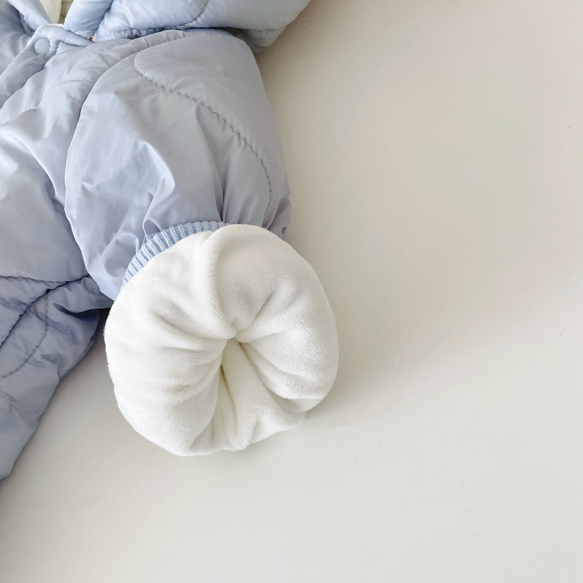 Close-up of a light blue padded jacket with a white, plush cuff, resting on a white surface. The jacket has visible stitching and is slightly crumpled at the elbow.