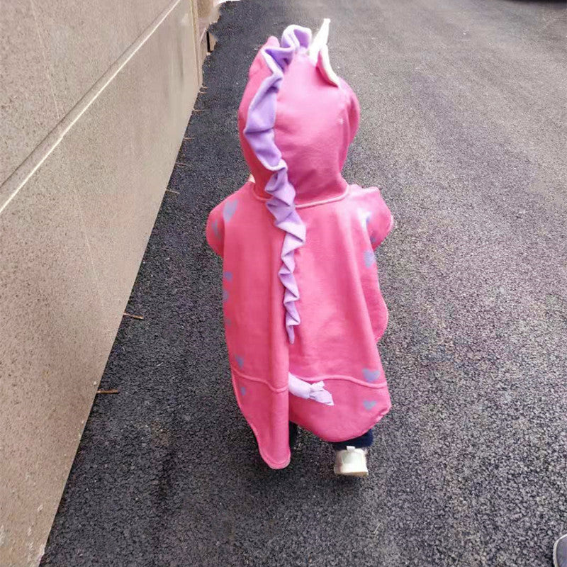Child in a pink dinosaur costume with purple spikes and a tail walks on a paved path, next to a concrete wall.