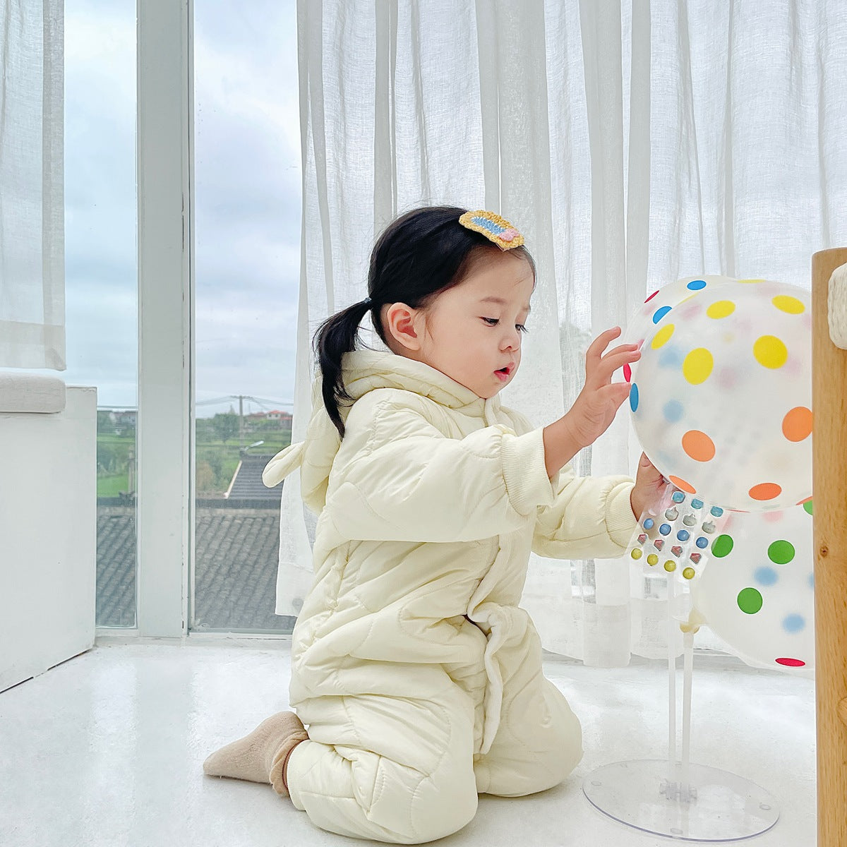 A toddler in a light cream snowsuit with a flower clip on her hair kneels by a window, interacting with a transparent balloon covered in colorful dots.